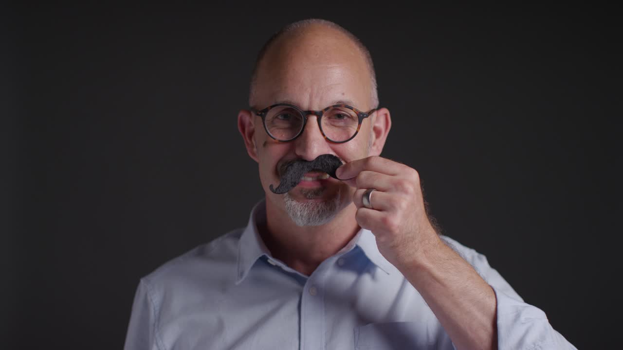 Portrait Of Mature Man Putting On Fake Moustache Symbolizing Support For Movember Promoting Men's Health And Cancer Awareness