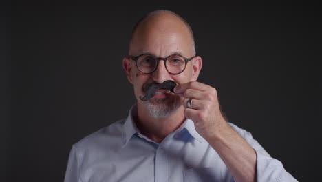 Portrait Of Mature Man Putting On Fake Moustache Symbolizing Support For Movember Promoting Men&#039;s Health And Cancer Awareness