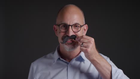 portrait of mature man putting on fake moustache symbolizing support for movember promoting men's health and cancer awareness