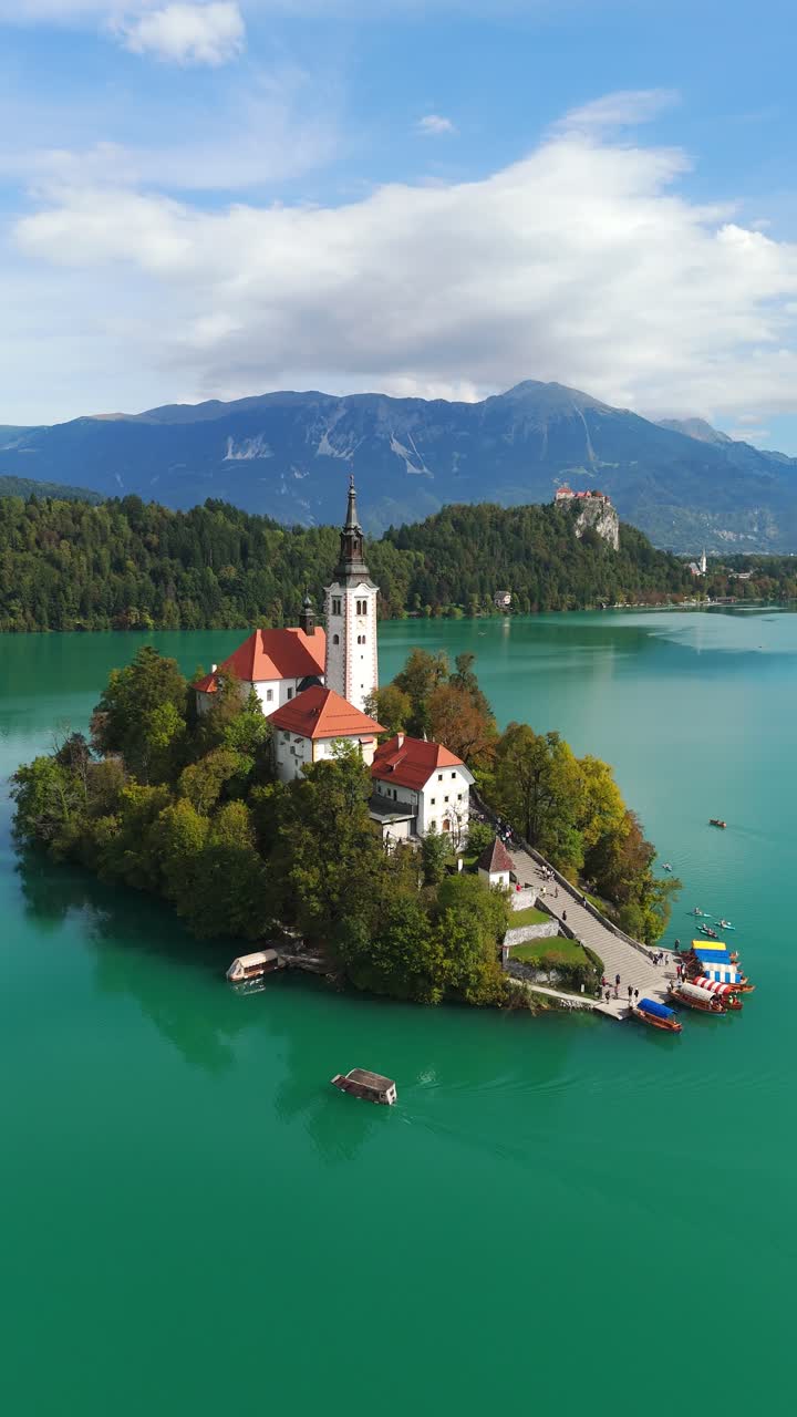Vista aerea verticale della pittoresca isola del lago di Bled con chiesa e vivace acqua turchese, il castello di Bled visibile sulla cima di una collina lontana sotto le Alpi Giulie in Slovenia
