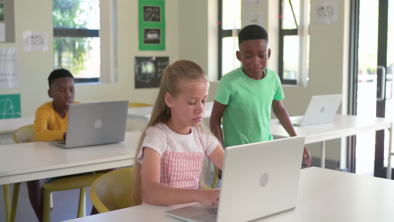Diverse child classmates watching green-shirt boy finishing laptop, walking to front desk, sharing