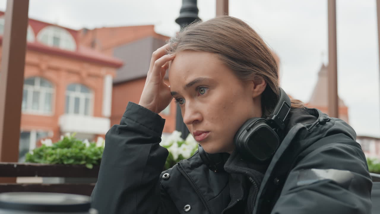 Женщина в наушниках пьет кофе из бумажного стакана в уличном кафе, снимает наушники и трет лоб, выражает обеспокоенность на фоне зданий из красного кирпича и пасмурного неба, на столе лежит смартфон