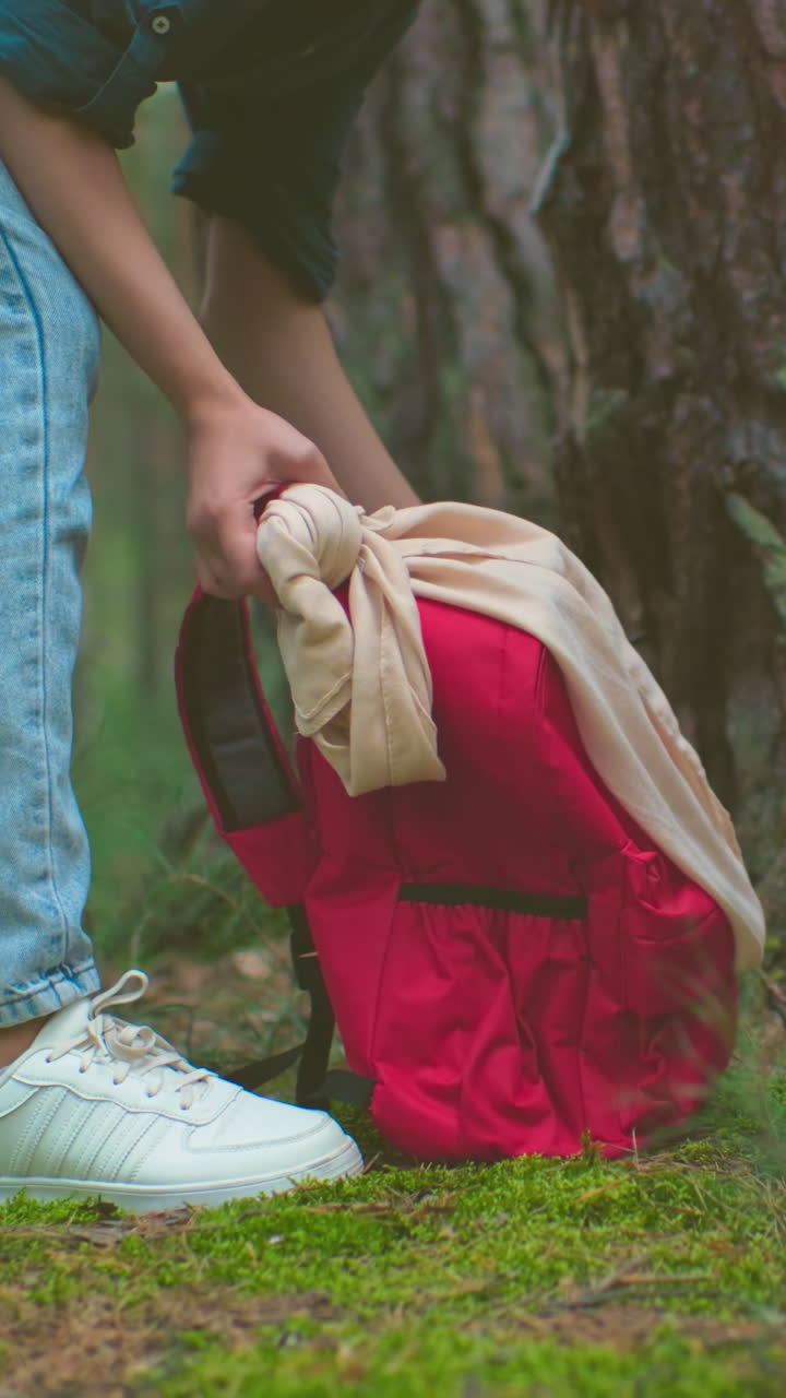 Young hikers retrieve their bags from ground in peaceful forest setting, with red bag tied with cloth, smiling woman in green shirt lifts bag over her shoulder, surrounded by lush trees and greenery