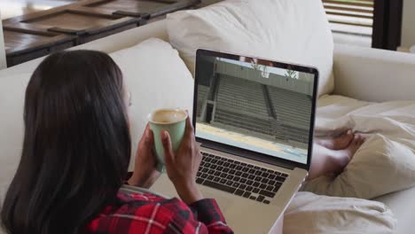 compuesto de una mujer sentada en casa sosteniendo café viendo un evento de salto de longitud de atletismo en una computadora portátil