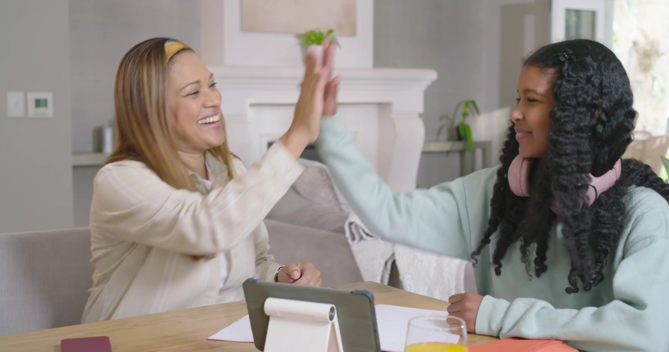 African American mother guiding child at table pointing tablet urging child writing in notebook
