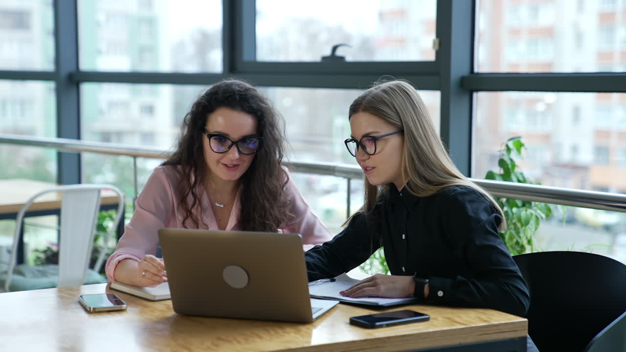 Two female office staff discussing something on the laptop screen. Business women working in the office atmosphere. Big windows at the backdrop.