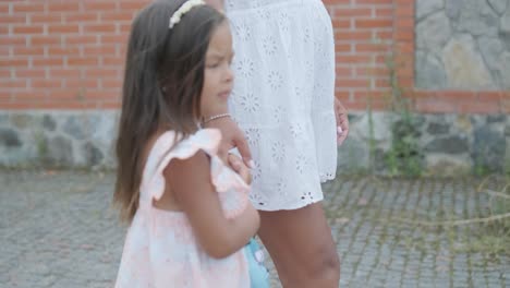 a pregnant woman and her young daughter, dressed in light summer outfits, walk hand-in-hand along a street in a residential neighborhood. the daughter holds a toy, enjoying the peaceful moment outdoor