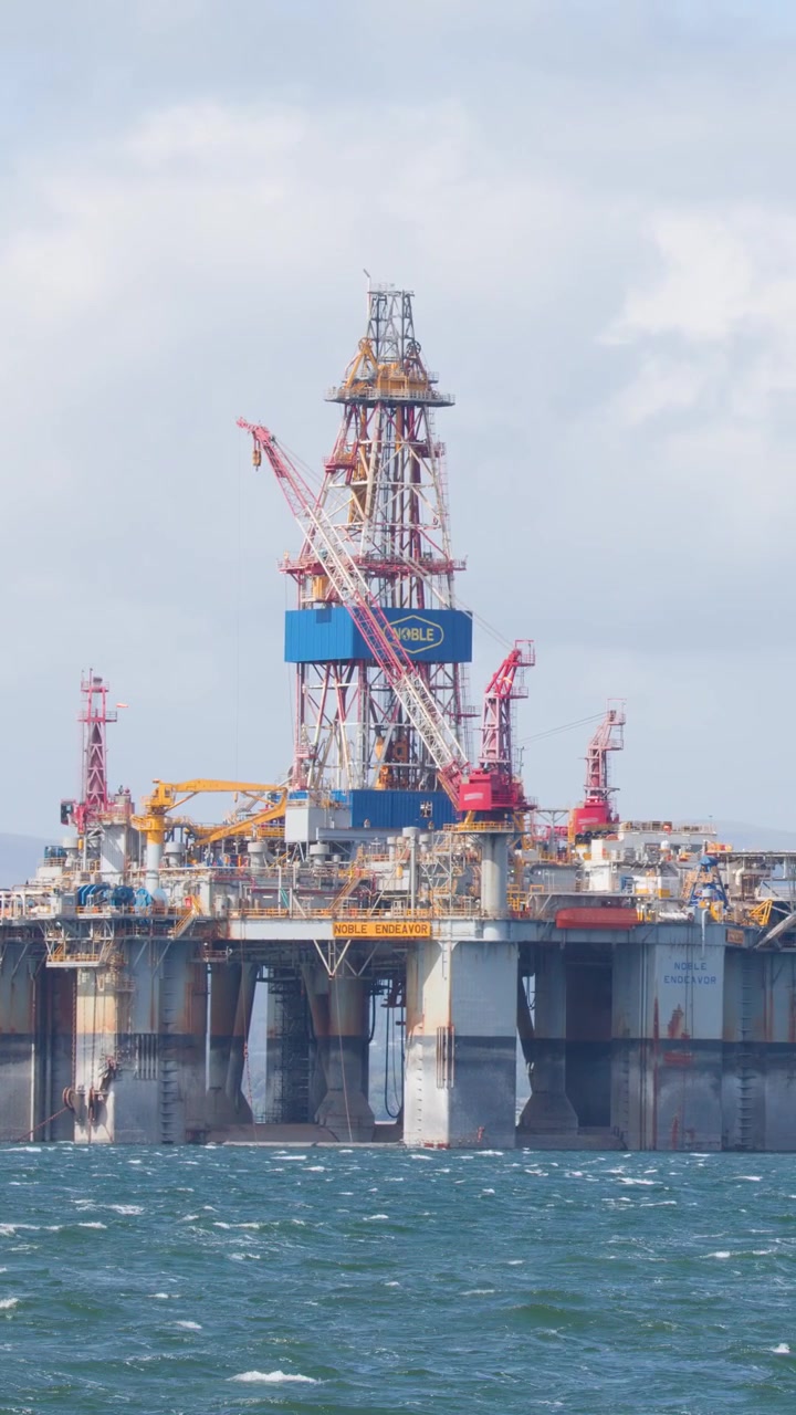 Stor offshore olieplatform i dagslys, blid kamerapanorering, oprørt hav, med De Skotske Højlande i baggrunden.