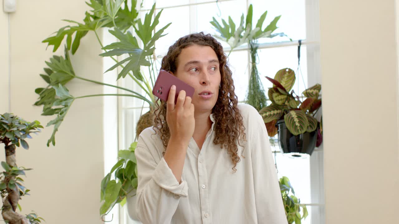 Nonbinary adult wearing cream top answering call holding purple phone by window plants chatting