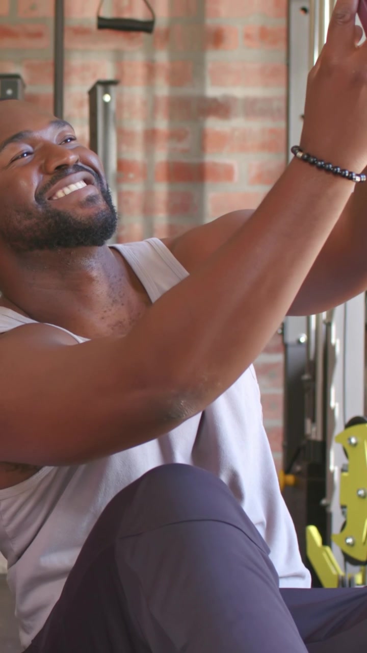 Vertical video: Adjusting man in tank angling maroon phone for selfie in gym brick wall bracelet