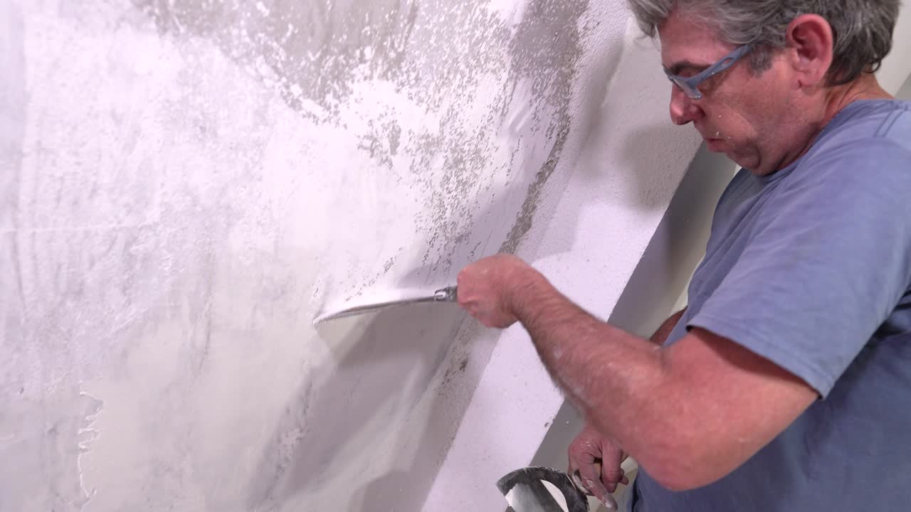 An adult plasterer is seen working on a new interior wall renovation, using a large metal trowel to evenly spread the white compound