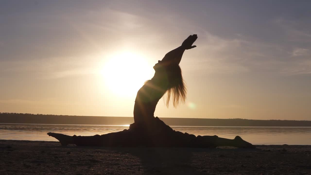 Silhouette of a beautiful yoga girl at sunrise on the beach. Hanumanasana, monkey pose. Sunrays lay on a body