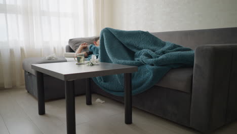 living room scene with boy lying on couch covered in blanket, glass cup and tissues on table with boy adjusting himself, coughing, and resting while feeling unwell