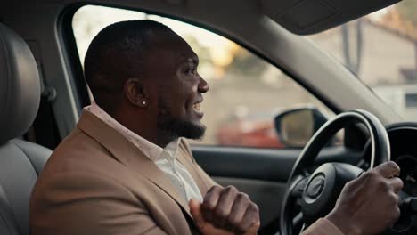 happy cheerful man businessman with black skin in a brown jacket drives a car while sitting in a modern car interior and dances during his business trip in the city