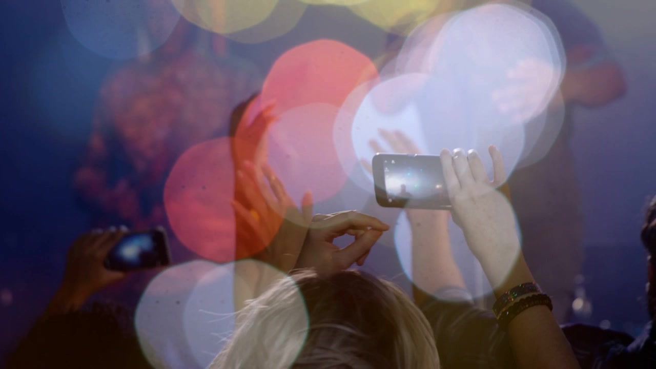 Video verticale: Fan al centro che alza il telefono filmando il palco a un concerto, con braccialetto e bokeh.
