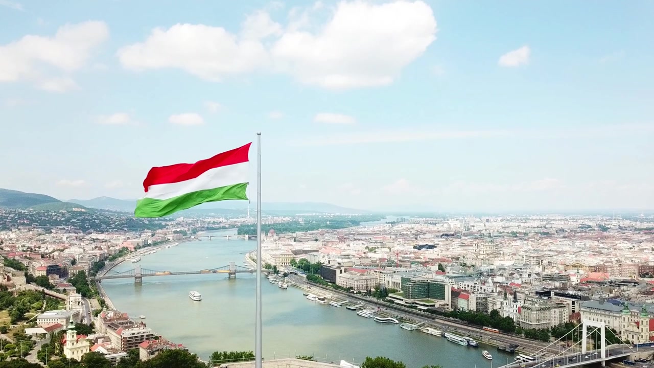 Bandiera ungherese che sventola fieramente al vento, dominando il panorama urbano di Budapest con il fiume Danubio che lo attraversa, sotto un cielo luminoso con nuvole bianche e soffici, ripresa aerea panoramica in slow motion.