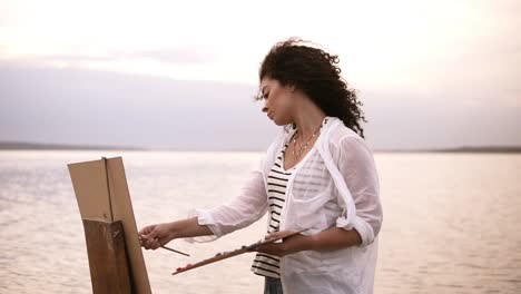 close up of an artist working process. an easel and holding palette in her hand. beautiful surrounding landscape: clear mirror lake and sky