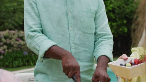 happy african american senior man cutting meat for lunch with friends in garden