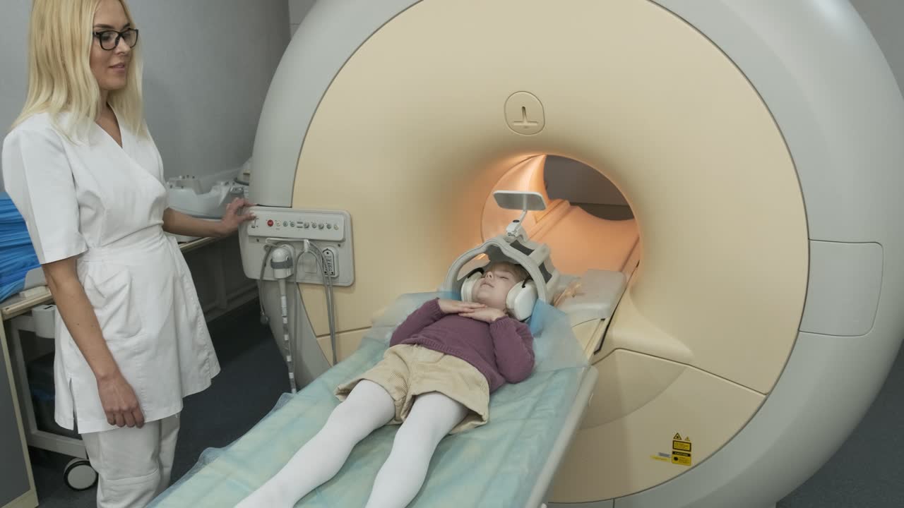 mujer médico radiólogo hace una resonancia magnética del cerebro, cabeza, cuello para el paciente niña pequeña, niña yace en la mesa automática, utilizando equipos modernos, bobina en la cabeza, auriculares