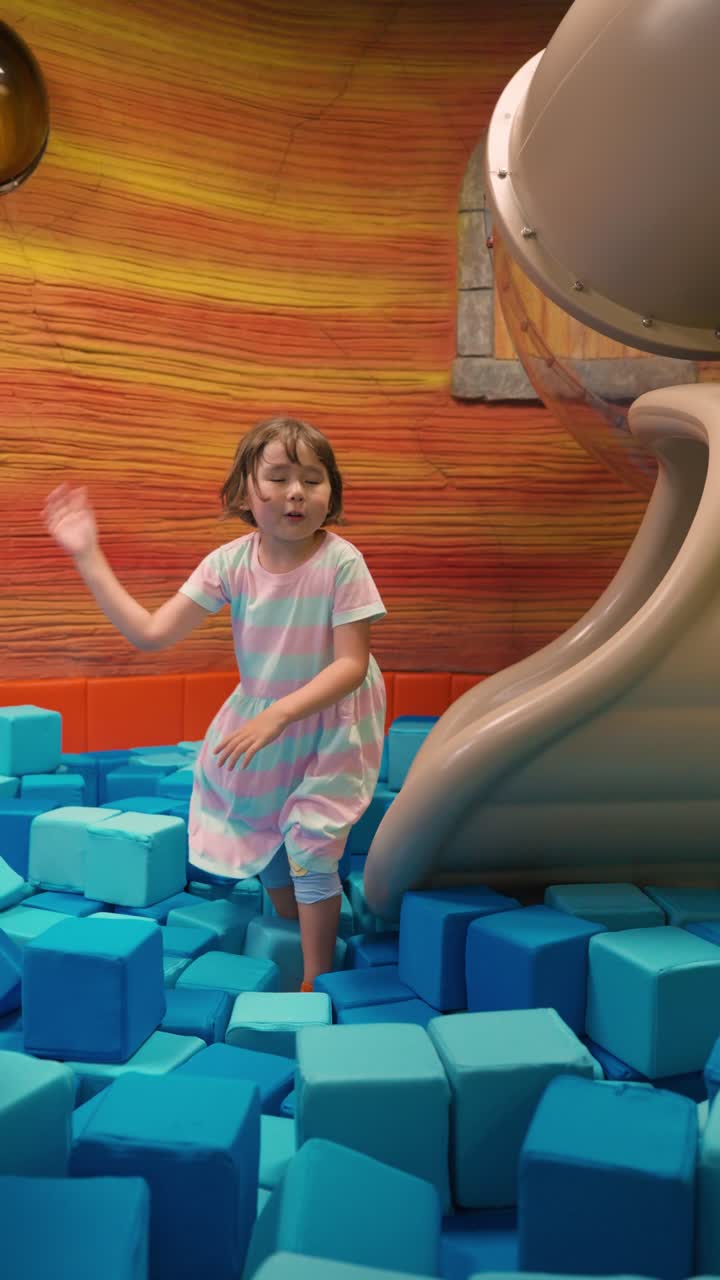 Happy five year old girl in a striped dress smiles as she lands at the bottom of a tube slide in a large pit filled with soft blue foam cubes