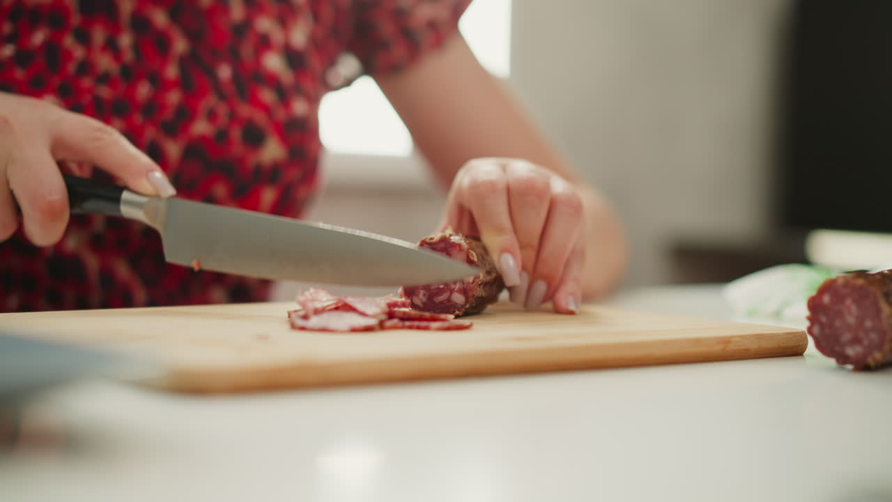 Elegant woman cutting thin slices of cured sausage on wooden board over white table by bright window light, highlighting precise knife skills, rich meat texture, home kitchen ambiance and gourmet snack prep