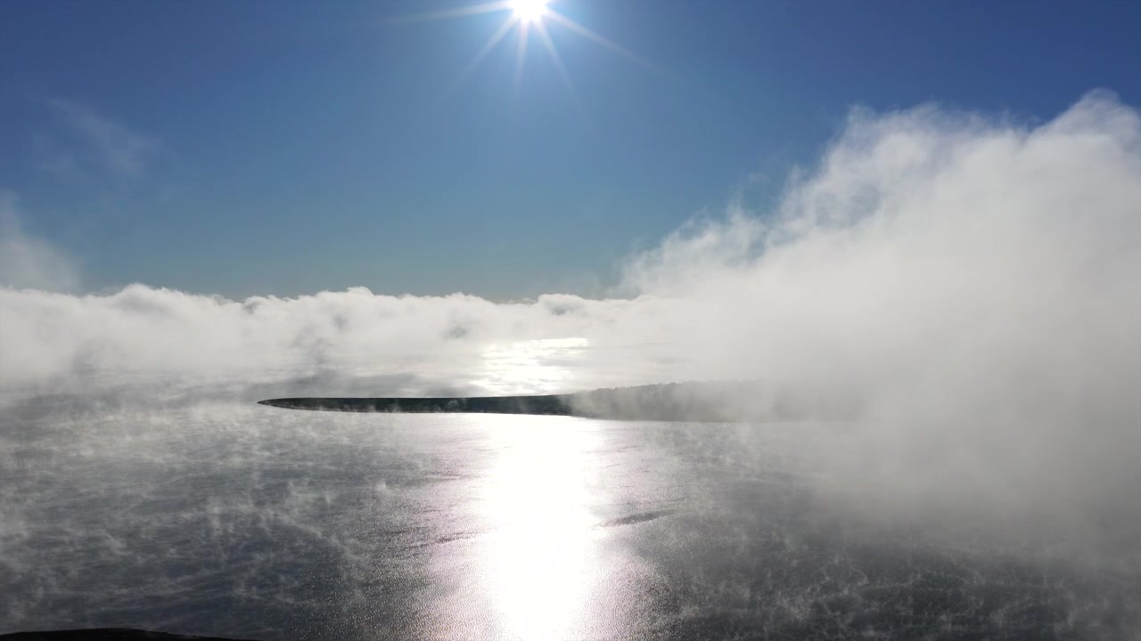 Morgenstrågetåge driver hen over et roligt ferskvandsreservoir, der reflekterer en lys solopgang, mens en skovklædt kystlinje dukker frem under en klar himmel, lav fremadgående droneflyvning.