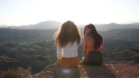 two young women sit on a cliff edge enjoying the desert mountain view at sunset