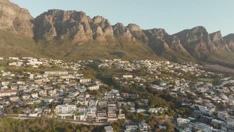 Drones-Volando-Alto-Hacia-Atrás-En-La-Cima-De-La-Bahía-De-Camps-En-Ciudad-Del-Cabo,-Sudáfrica---Muchas-Casas-En-Una-Pendiente---Vista-De-La-Montaña-De-La-Mesa-Y-El-Amplio-Mar-Al-Atardecer