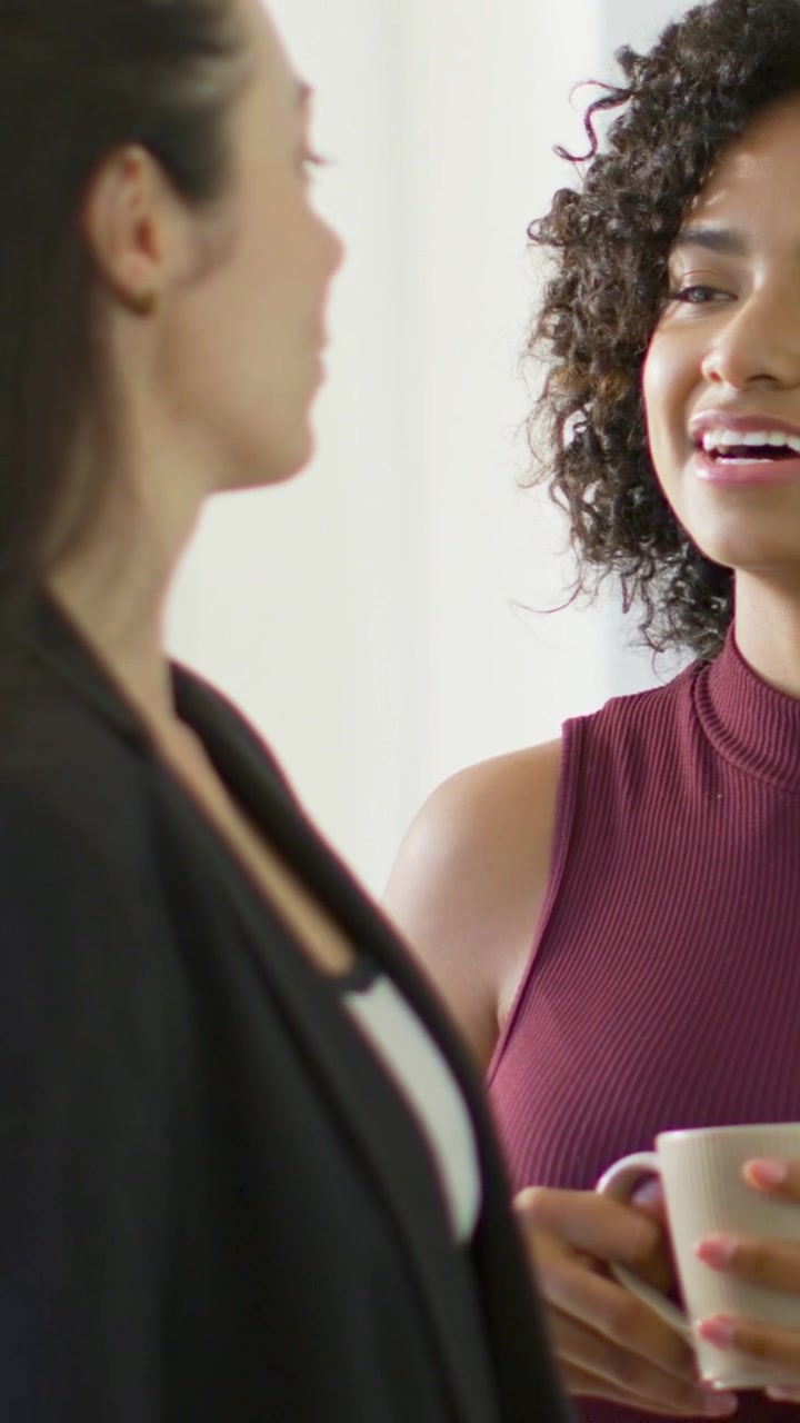 Vídeo vertical: Mulher sorridente à direita iniciando uma conversa no intervalo enquanto segura uma caneca branca, com roupas de trabalho.