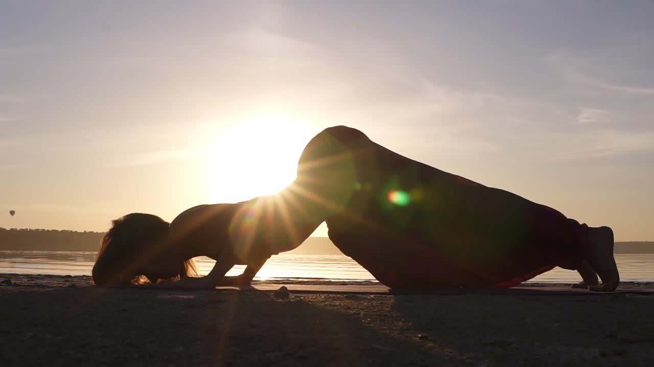 A silhouette footage of a woman who is doing Urdhva Mukha Shvansana on the beach. Backbend. Sun shines on the background