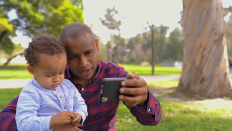 Father-sitting-on-grass,-holding-son-on-knees,-making-selfie