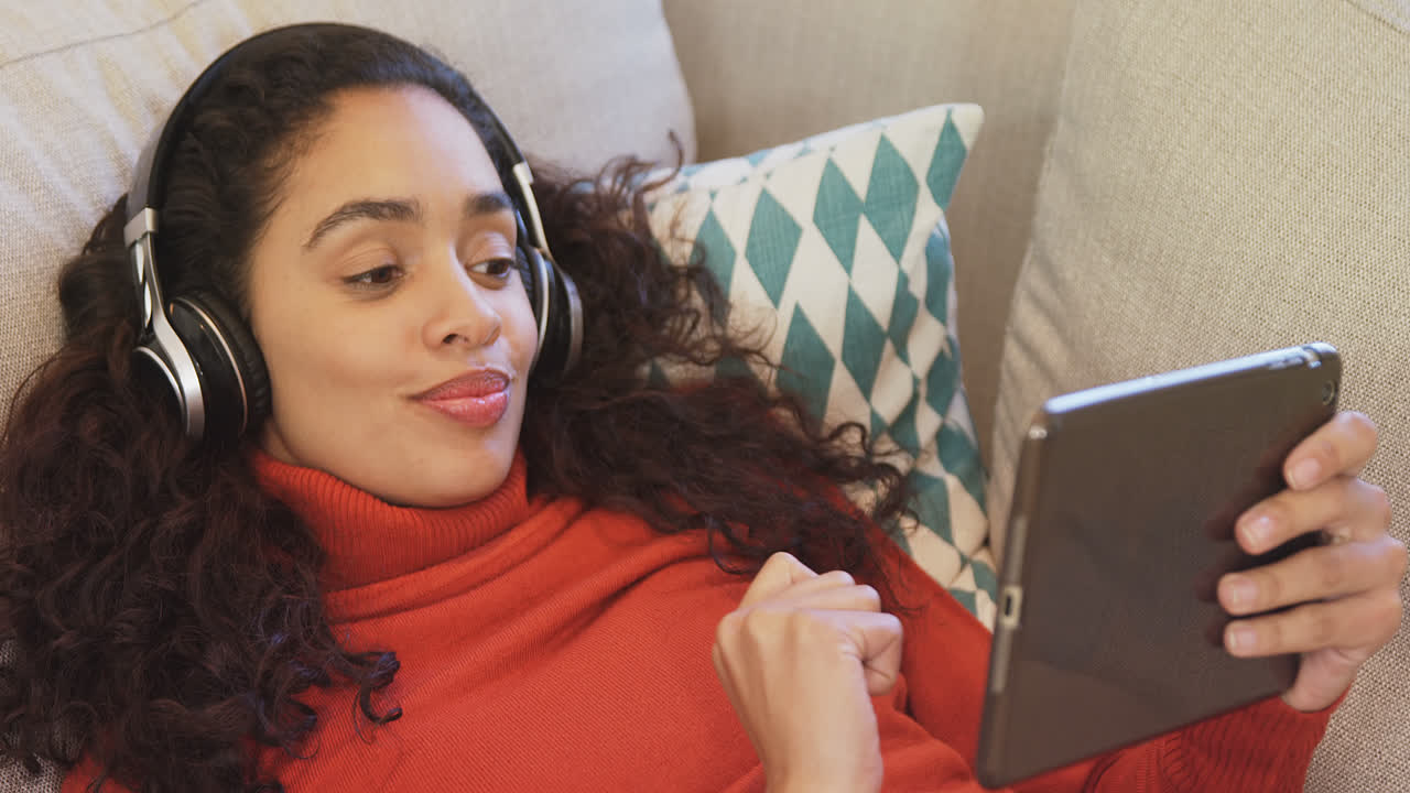 Happy woman lying on sofa using tablet enjoying music on her headphones 4K 4k