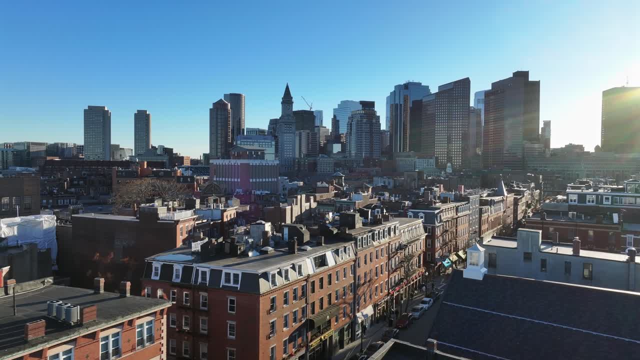 Bustling Boston skyline rises behind North End rowhouses. Custom House tower stands tall among downtown skyscrapers. Aerial during sunset.