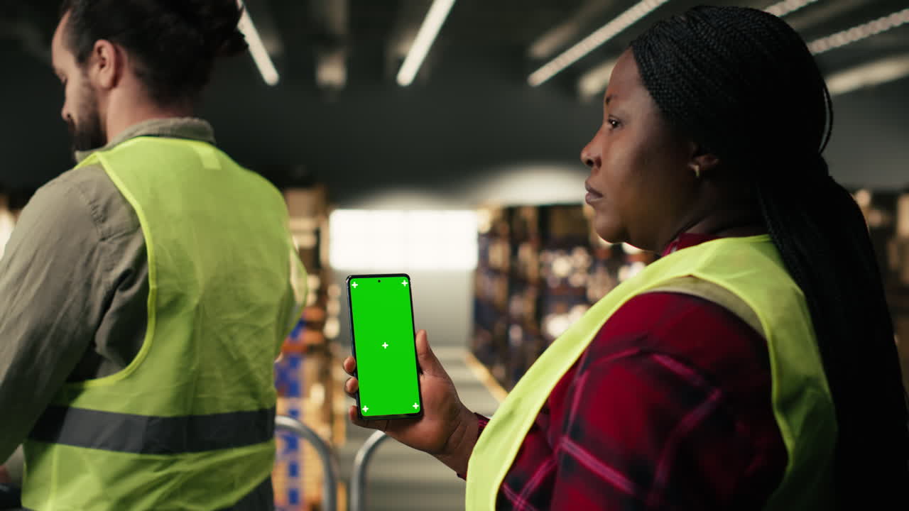 Warehouse Worker Holding Green Screen Smartphone While Colleague Works on Computer