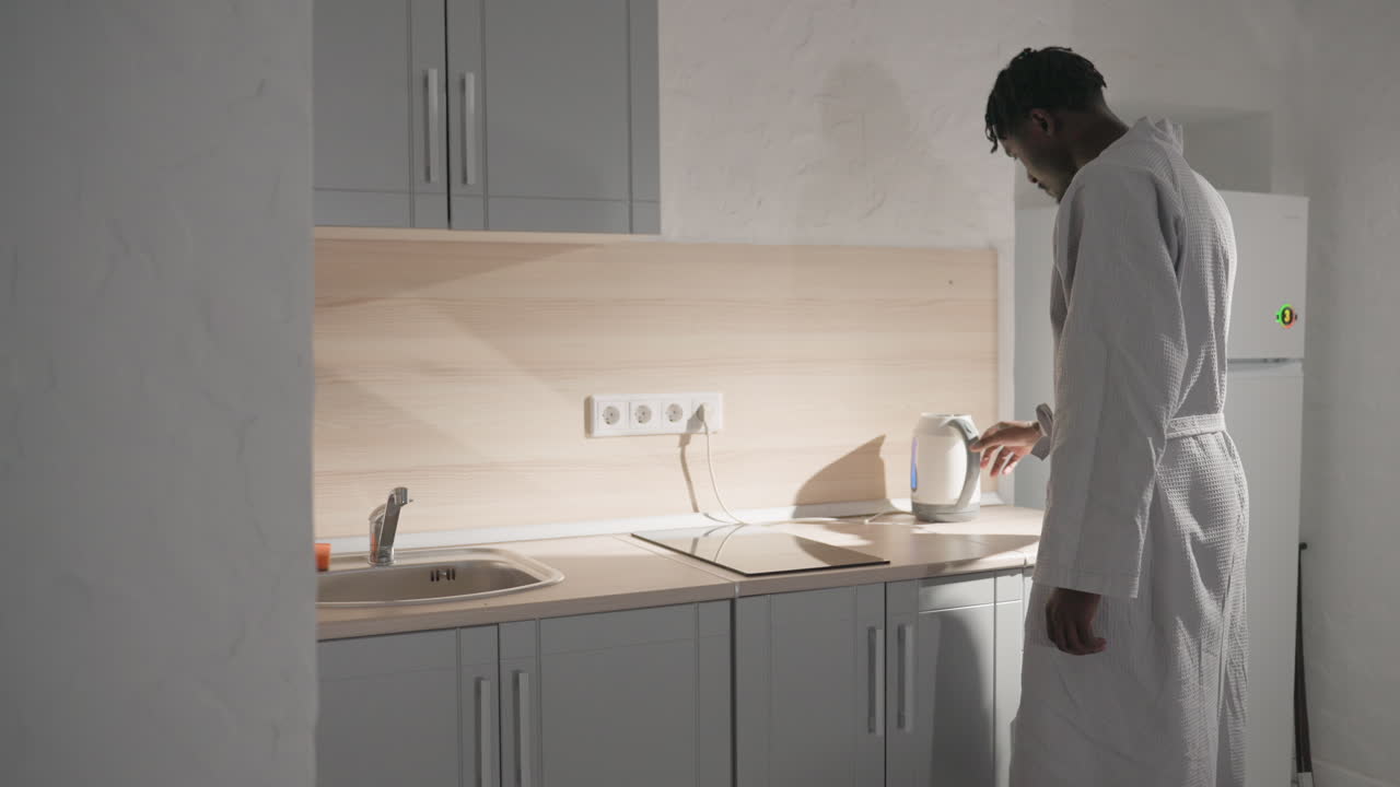 Guy in bathrobe standing in kitchen, filling kettle with water. Soft light illuminating modern kitchen with clean countertops. Relaxed, peaceful morning routine in cozy indoor space