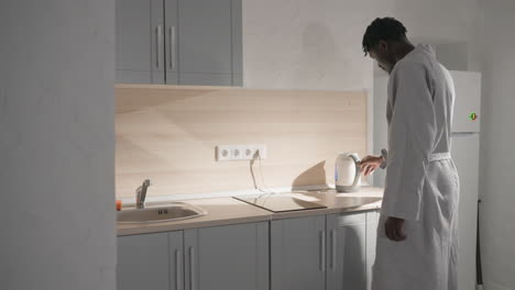 guy in bathrobe standing in kitchen, filling kettle with water. soft light illuminating modern kitchen with clean countertops. relaxed, peaceful morning routine in cozy indoor space