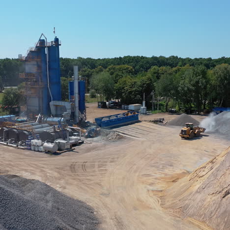 industrial territory surrounded by nature in summer. asphalt plant and piles of sand and rubble outdoors. excavator transports gravel. aerial view.