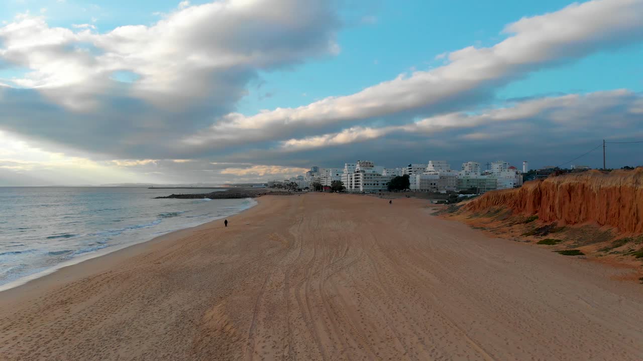 playa con la ciudad de quarteira al fondo, rodeada por un acantilado, un mar en calma y un cielo nublado