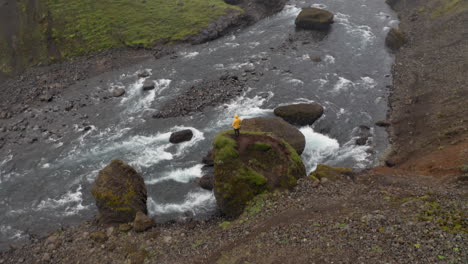 aerial: panning shot of a man wearing yellow jacket, standing on a rock near a river