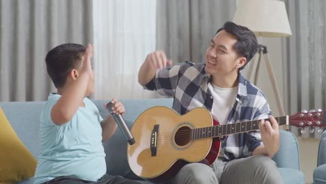 happy asian father and son celebrating for being able to play the guitar and singing together on sofa at home