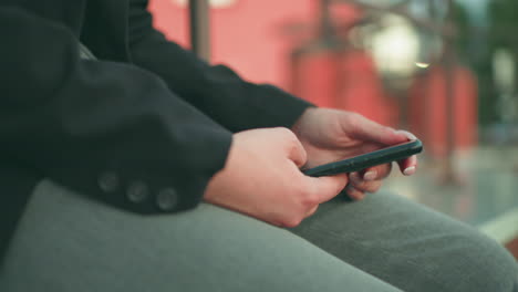 side view of lady with artificial nails holding smartphone and operating device while seated in urban environment, dressed in black blazer, with bokeh lights and buildings in soft background