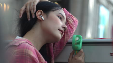 Asiatic-woman-listening-to-music-with-wireless-earphones-refreshing-cool-air-using-mini-portable-usb-fan-facing-the-summer-heat-due-to-global-warming