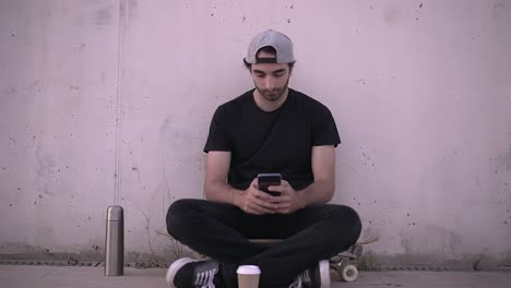 young man taking a selfie with a coffee cup while sitting on a skateboard.