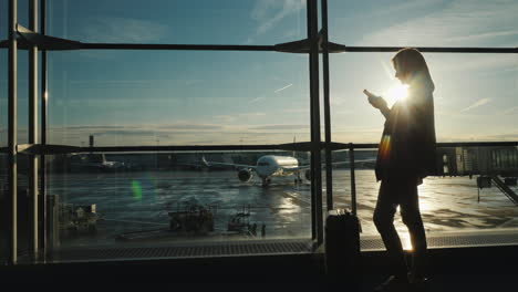 Pasajero-Con-Un-Teléfono-Inteligente-En-La-Terminal-Del-Aeropuerto,-La-Silueta-Ilumina-El-Sol-Detrás-De-La