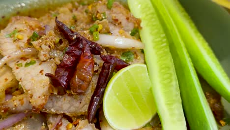 a close-up of spicy pork topped with dried chili, lime slices, and fresh cucumber sticks.