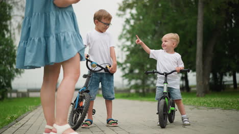 two siblings are seen interacting joyfully, with the younger one pointing at his older brother as they both enjoy stand on the bicycle, a woman, seen from behind, stands nearby