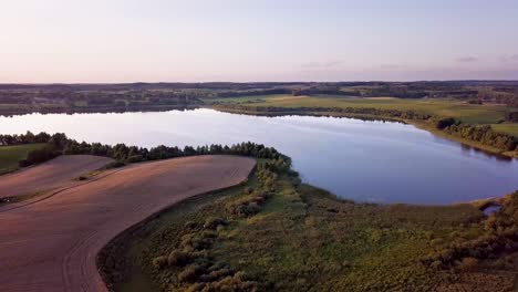 Toma-Aérea-De-Un-Lago-Pequeño-Y-Tranquilo-Rodeado-De-árboles-Y-Granjas-Al-Atardecer