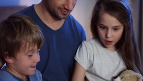 close-up view of caucasian father with two kids sitting on the bed at night, talking and looking at something