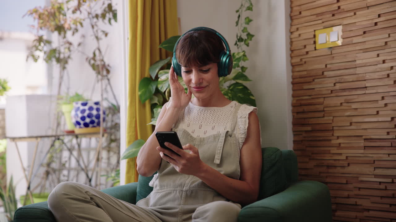 Donna che usa lo smartphone mentre ascolta musica con le cuffie a casa. Momento di vita quotidiana casuale, piacere e interazione digitale quotidiana in un ambiente interno accogliente