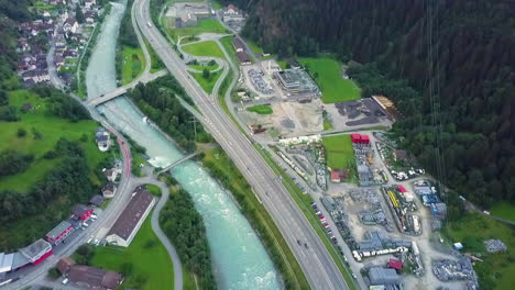 Vehículos-Que-Conducen-Por-La-Carretera-A-Lo-Largo-Del-Río-Y-La-Ciudad-Con-Vistas-Panorámicas-De-Las-Montañas-Verdes-En-Verano-En-Logano,-Suiza